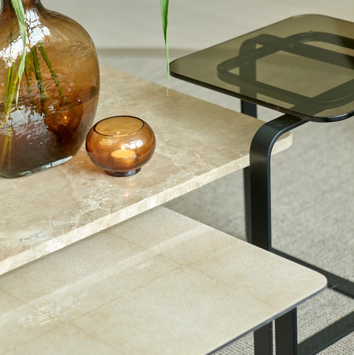 Close-up of stacked Lora coffee tables with beige stone, leather, and smoked glass tops, decorated with amber glass vases.