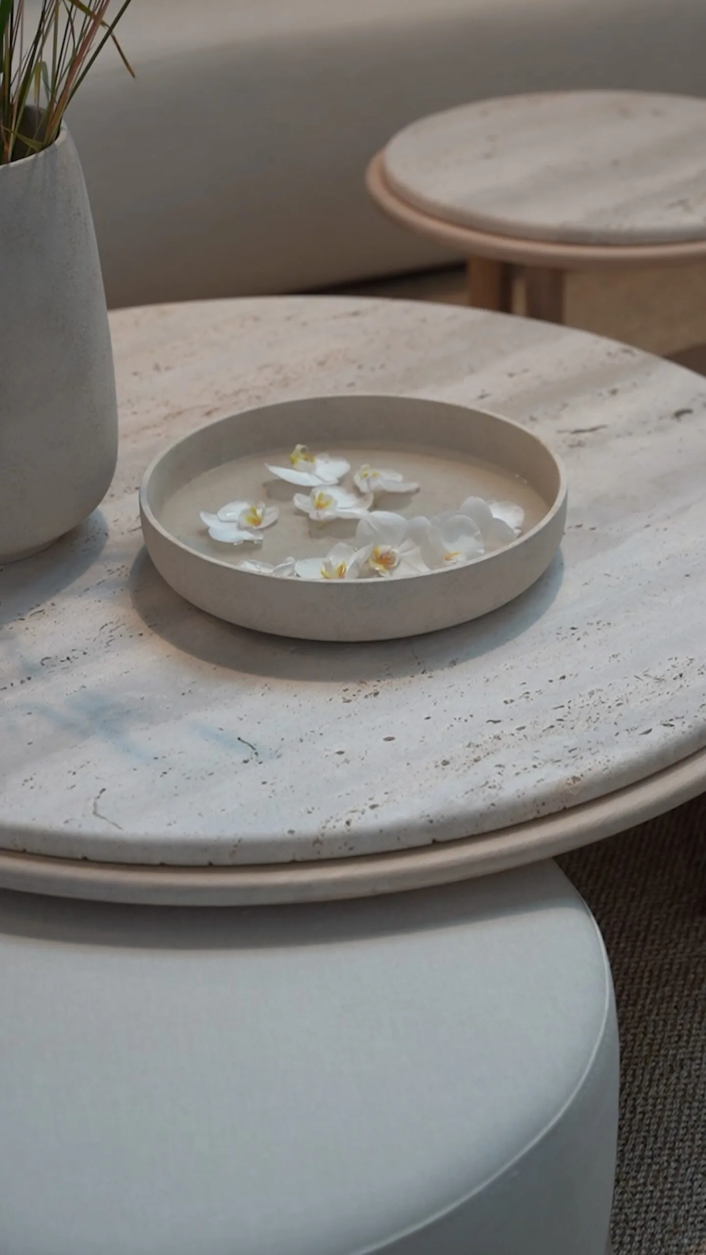 Round beige Bola bowl with floating white orchids on a light travertine coffee table.