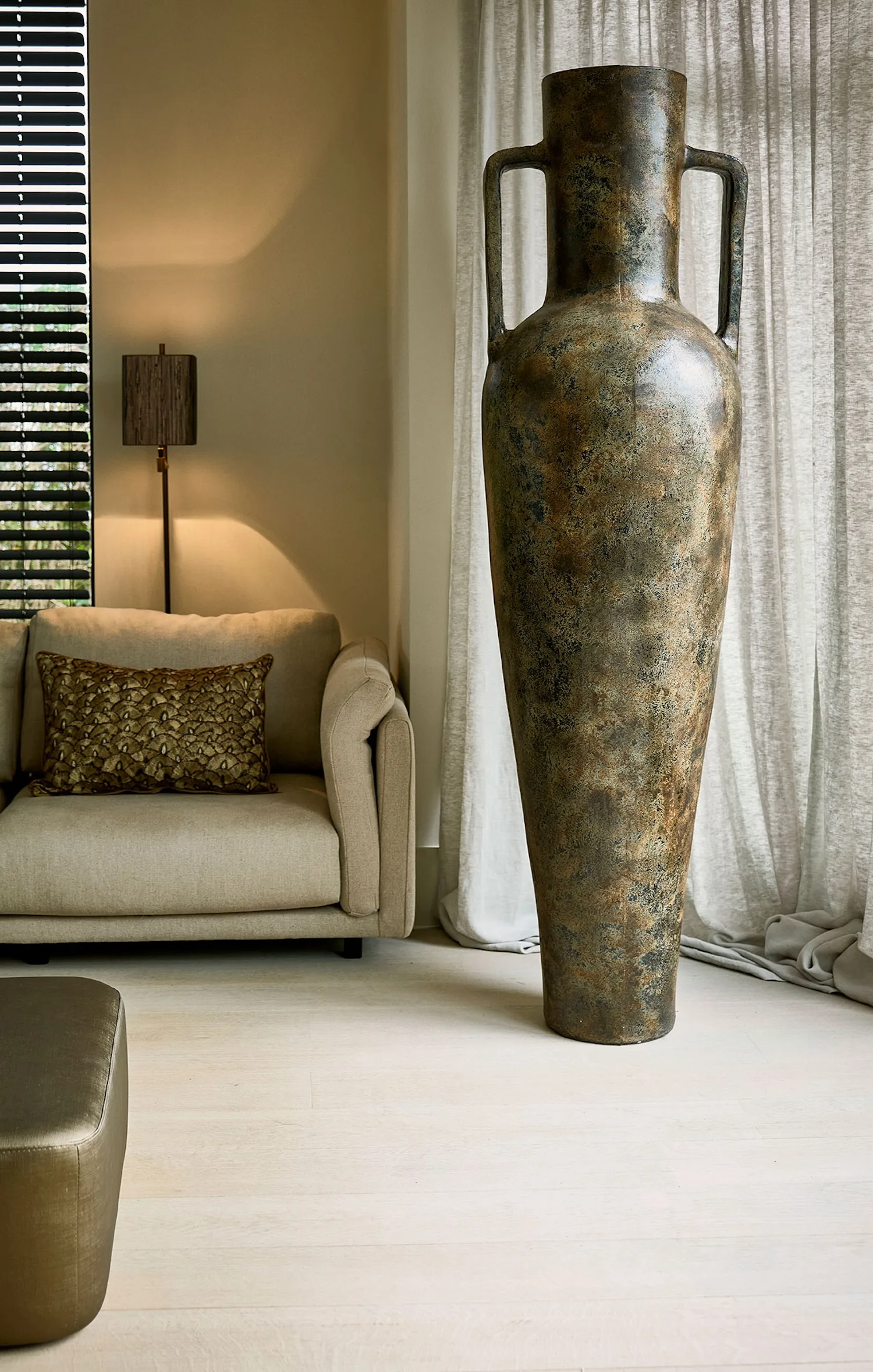 Tall bronze-toned Vorera vase with side handles, placed beside a beige sofa and floor lamp in a modern interior.