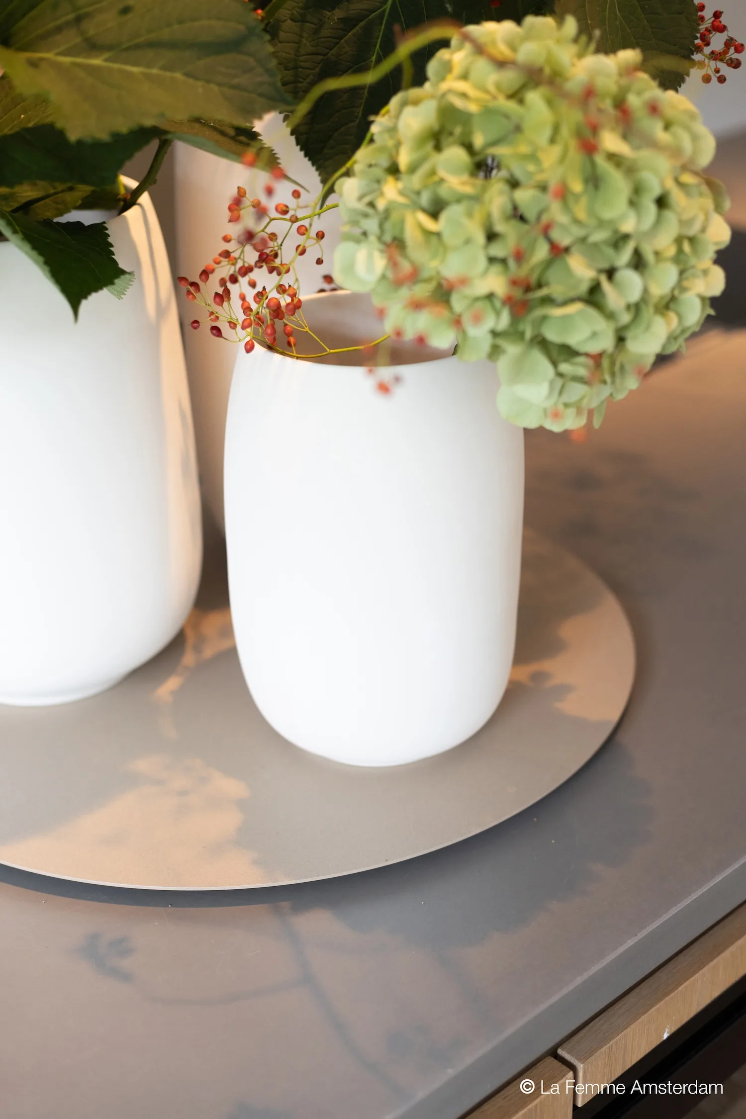 Close-up of a white Bola vase with green hydrangea and red berries on a grey surface.