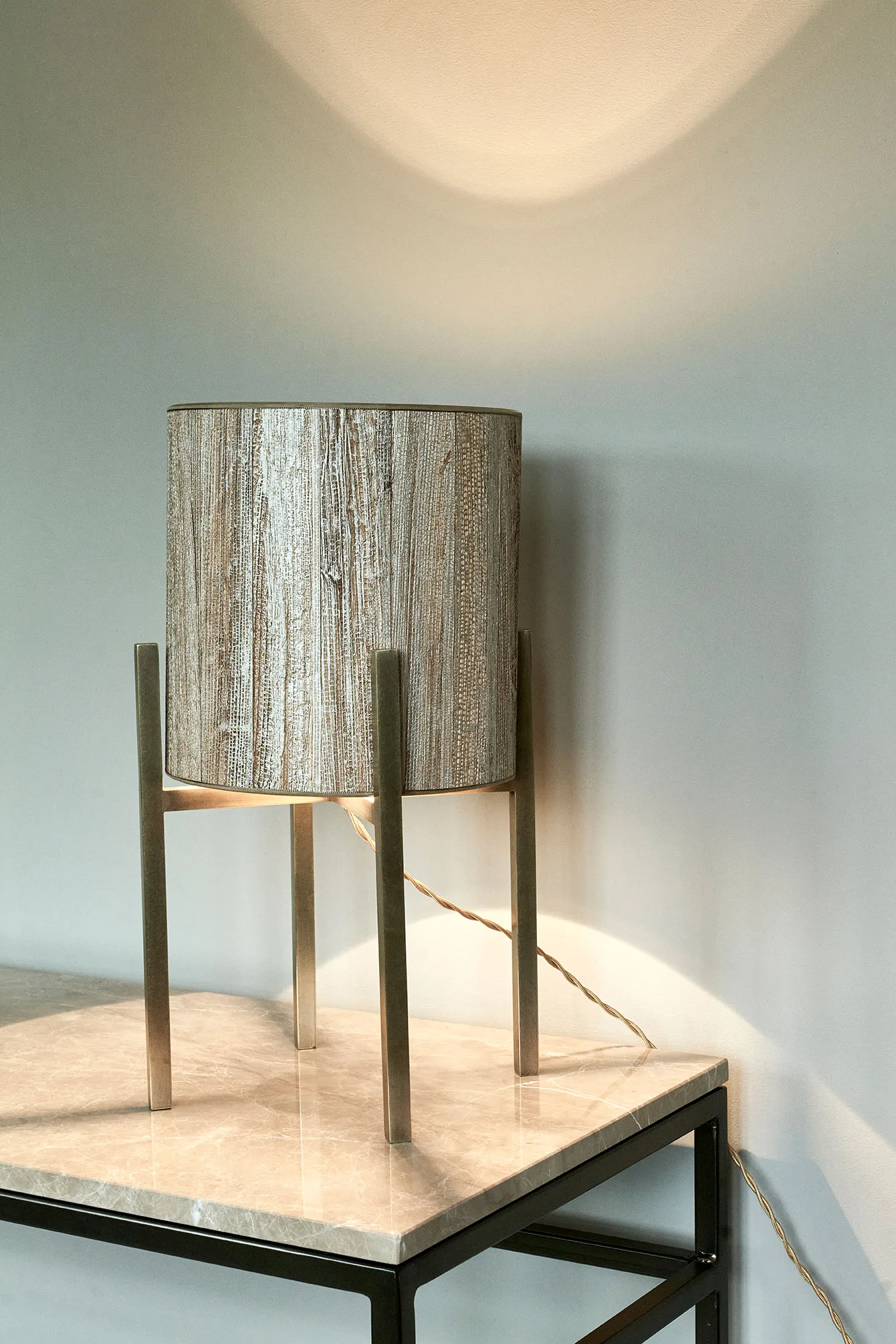 Cylindrical lamp with textured shade and four-legged metal base, placed on a stone tabletop.