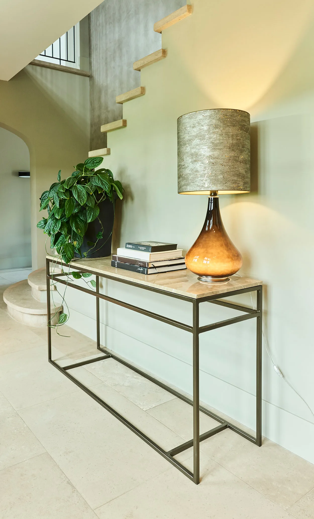 Console styled in an entryway with a table lamp, books, and a plant beside stairs.