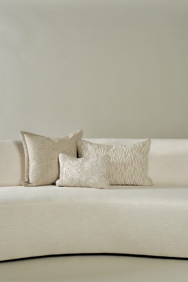 Three beige-toned decorative cushions on a light curved sofa against a neutral wall.