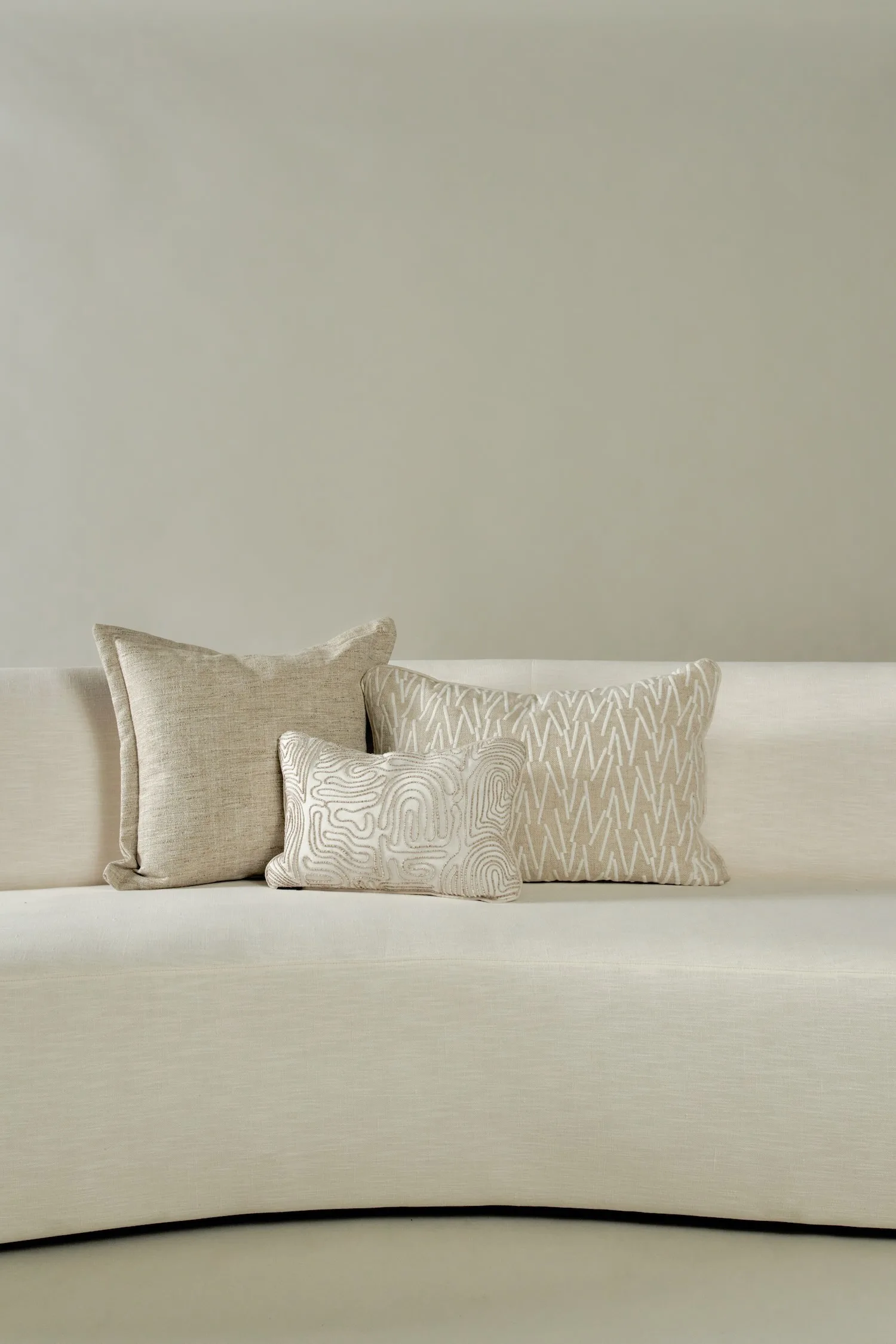 Three beige-toned decorative cushions on a light curved sofa against a neutral wall.