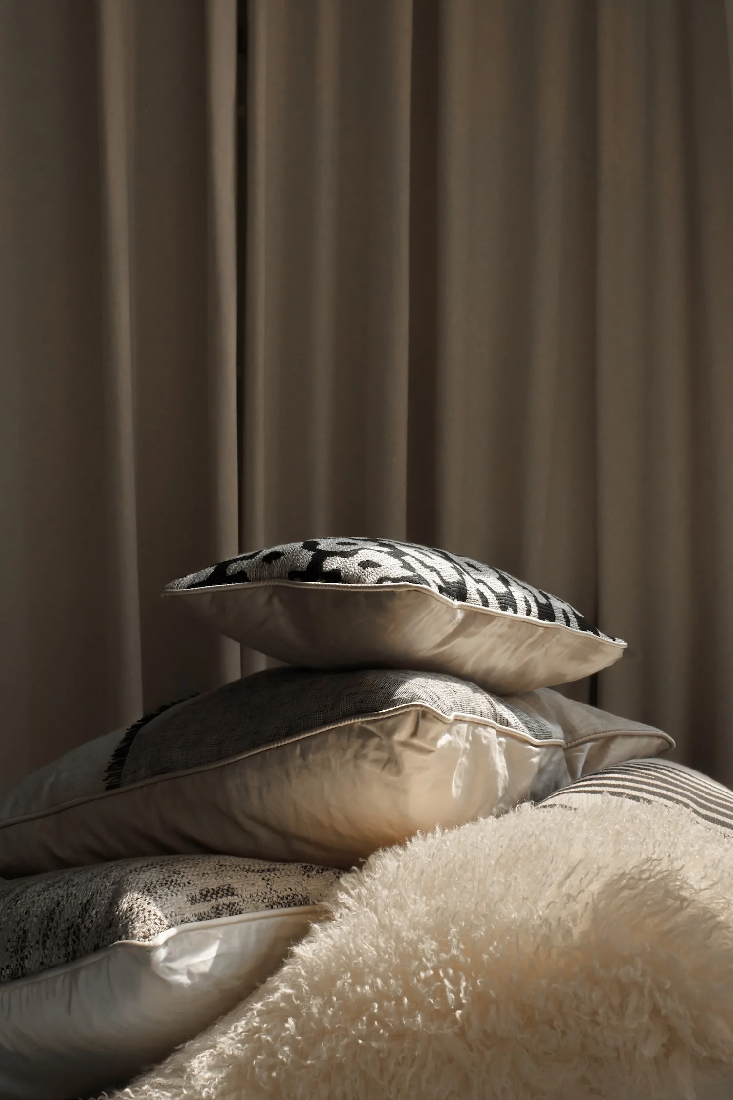Stacked cushions in beige, grey, and black tones with soft and woven textures, set against neutral curtains.