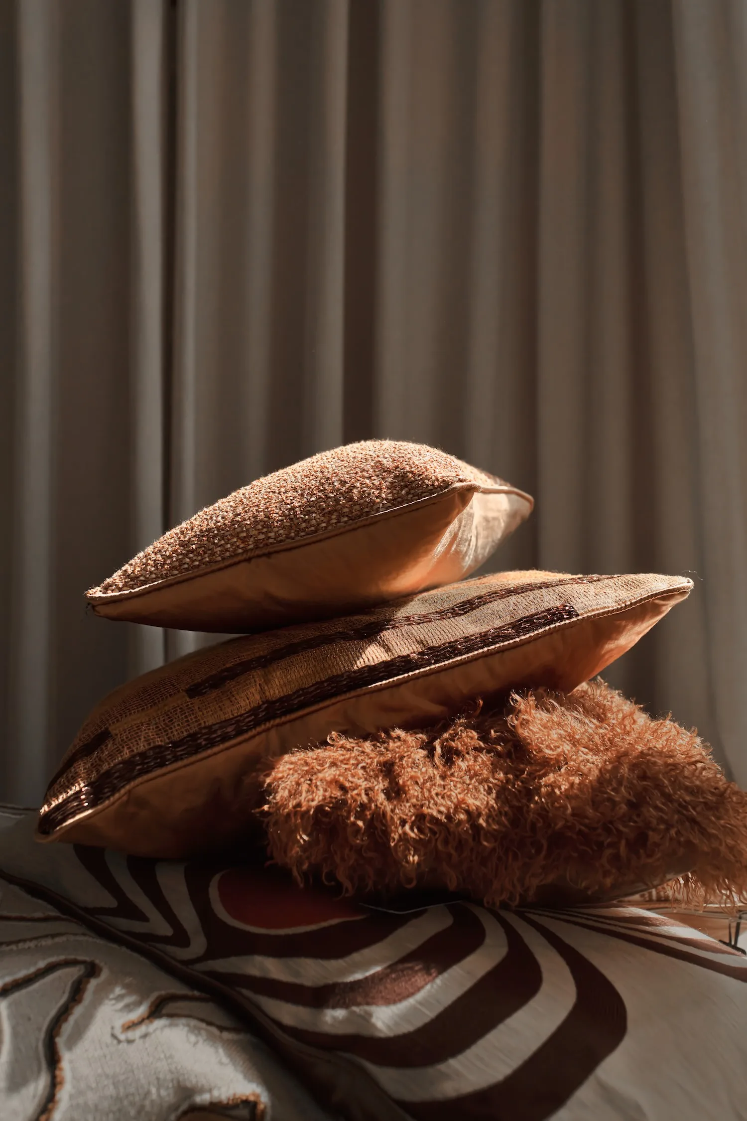 Close-up of stacked cushions in bronze, terracotta, and brown hues with varying textures and fabrics.