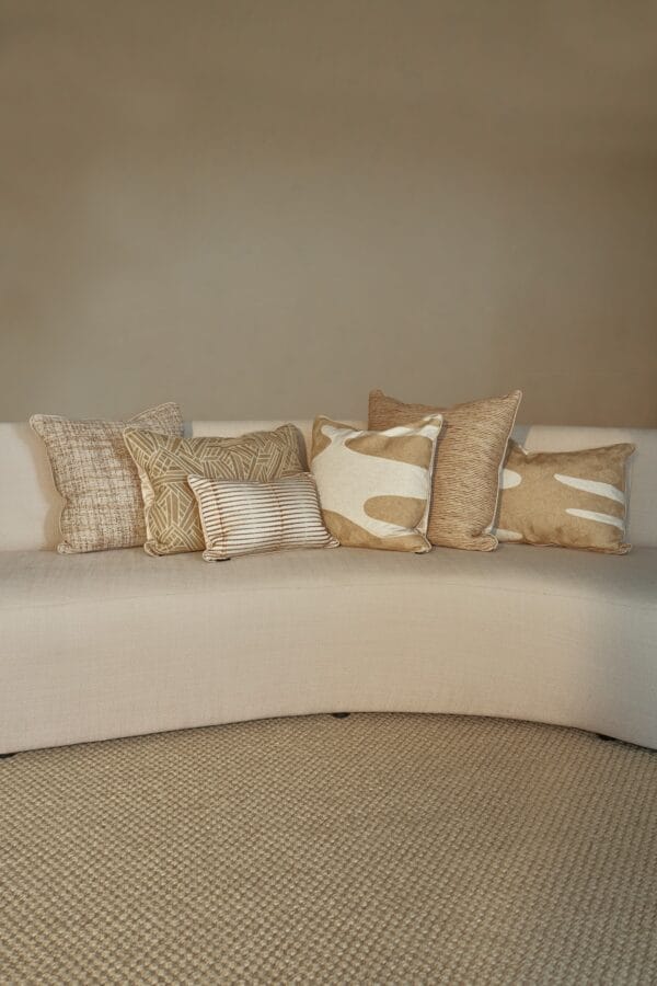 Assortment of six textured and patterned cushions in warm earthy tones on a light beige curved sofa.