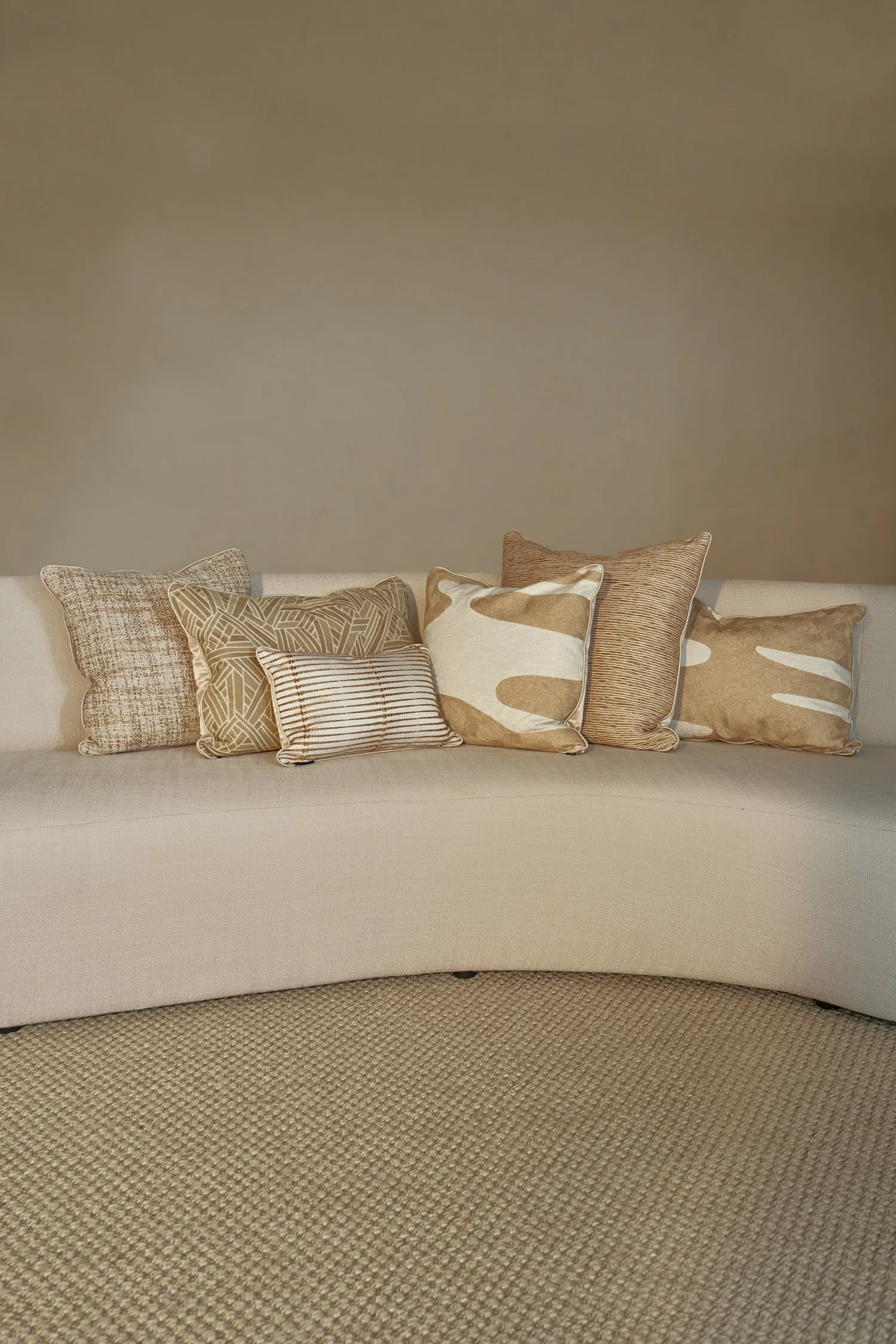 Assortment of six textured and patterned cushions in warm earthy tones on a light beige curved sofa.