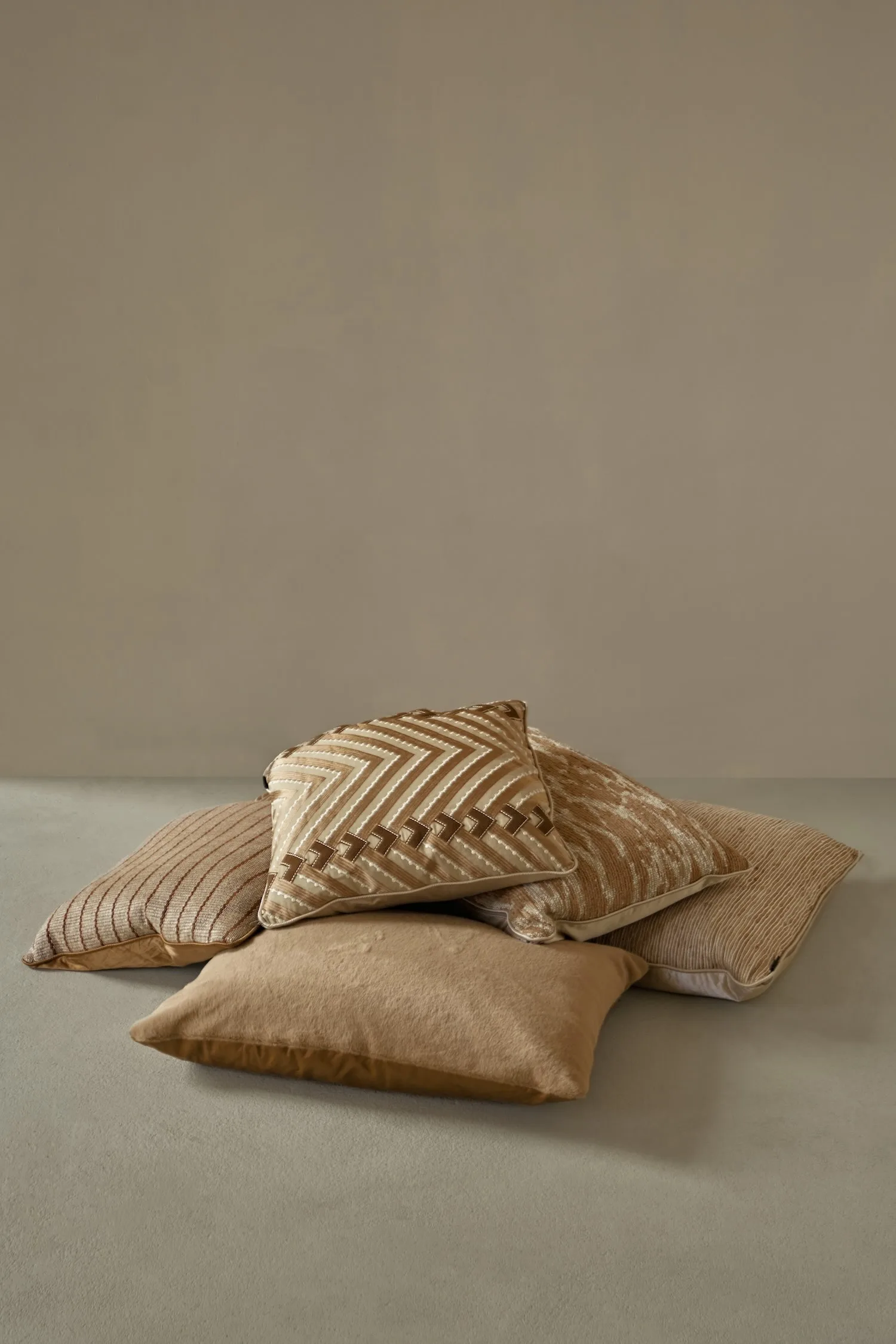 Five decorative cushions in rich brown and beige tones stacked casually on a smooth grey floor.