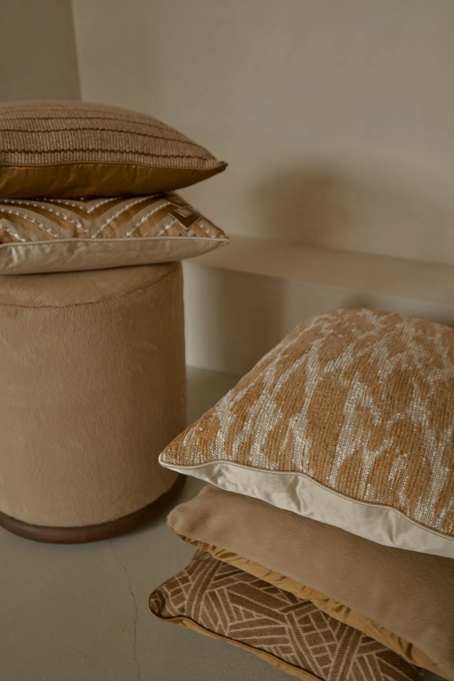 Decorative cushions with textured and geometric patterns stacked beside a soft brown pouf.