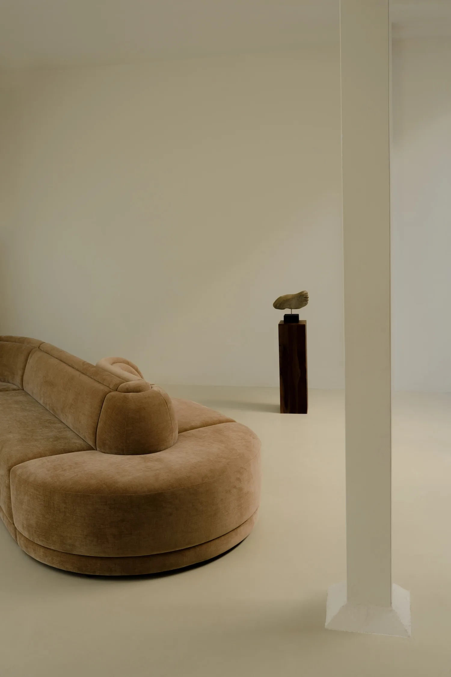 Curved Georgie modular sofa in camel velvet fabric placed in a minimalist interior with a sculptural piece on a pedestal in the background.