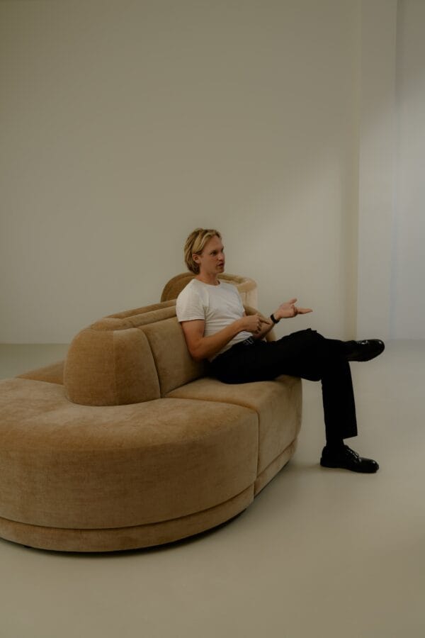 Maurits sitting on a large camel-colored Georgie modular sofa in a minimalist, softly lit room, engaged in conversation.