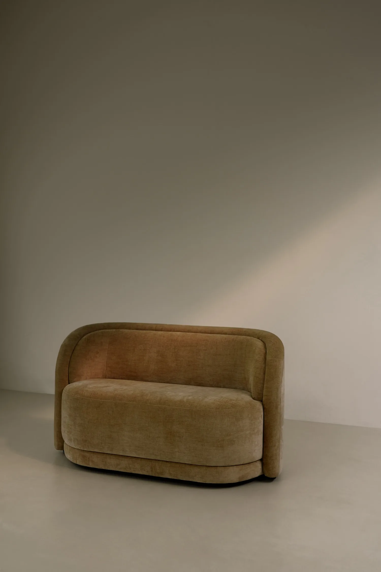 Compact two-seater camel-colored Georgie sofa with soft curves, placed against a neutral wall with a shadow of light.