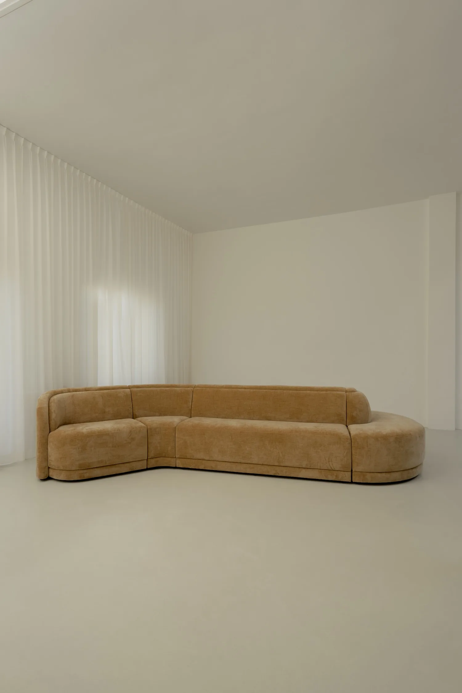 Wide-angle view of a camel-colored L-shaped Georgie modular sofa in a minimalist living room with soft white curtains and neutral walls.