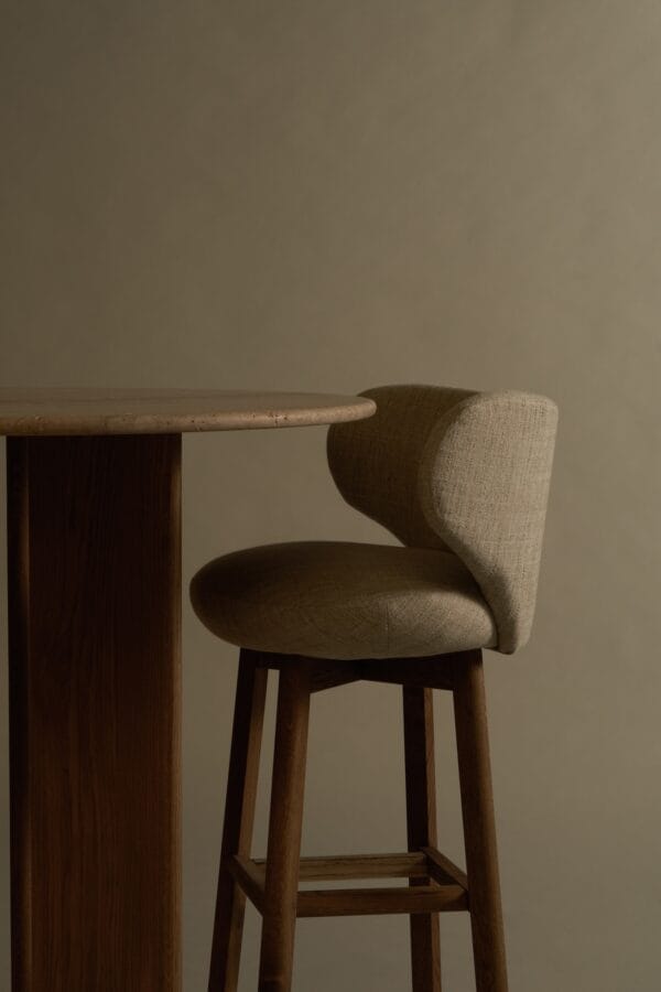 Margaux barstool in light fabric next to a wooden bar table, low light setting.