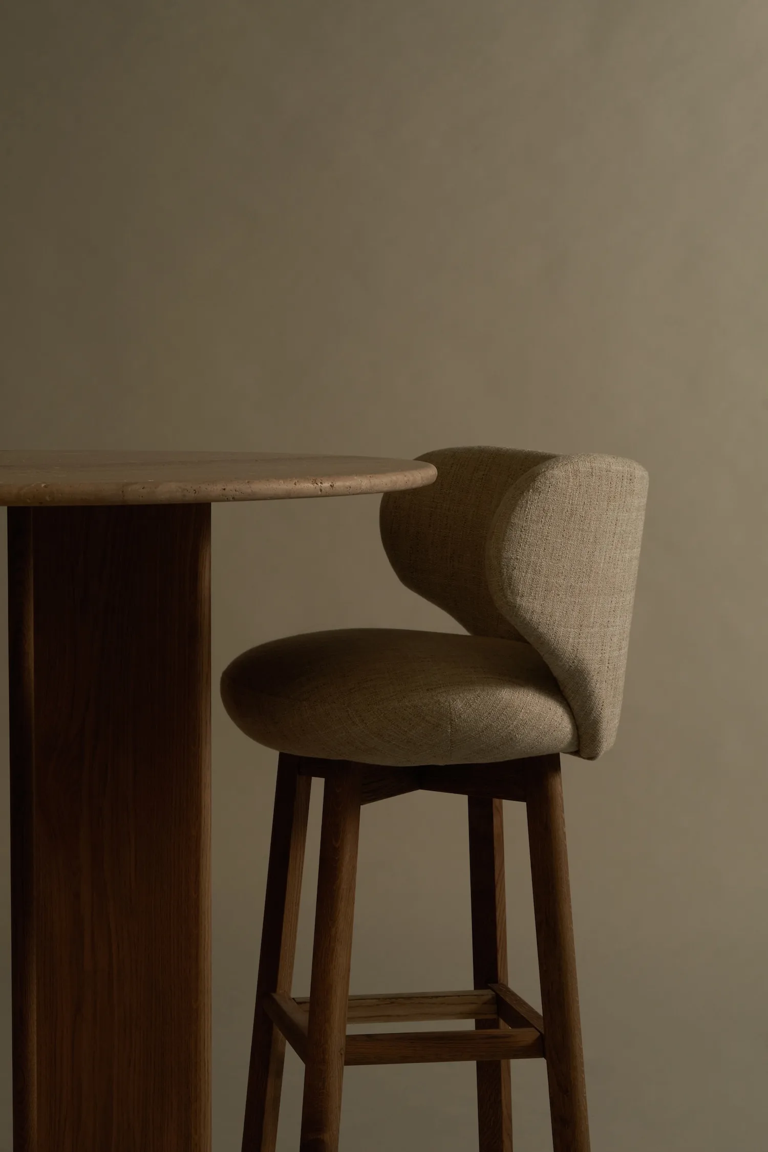 Margaux barstool in light fabric next to a wooden bar table, low light setting.