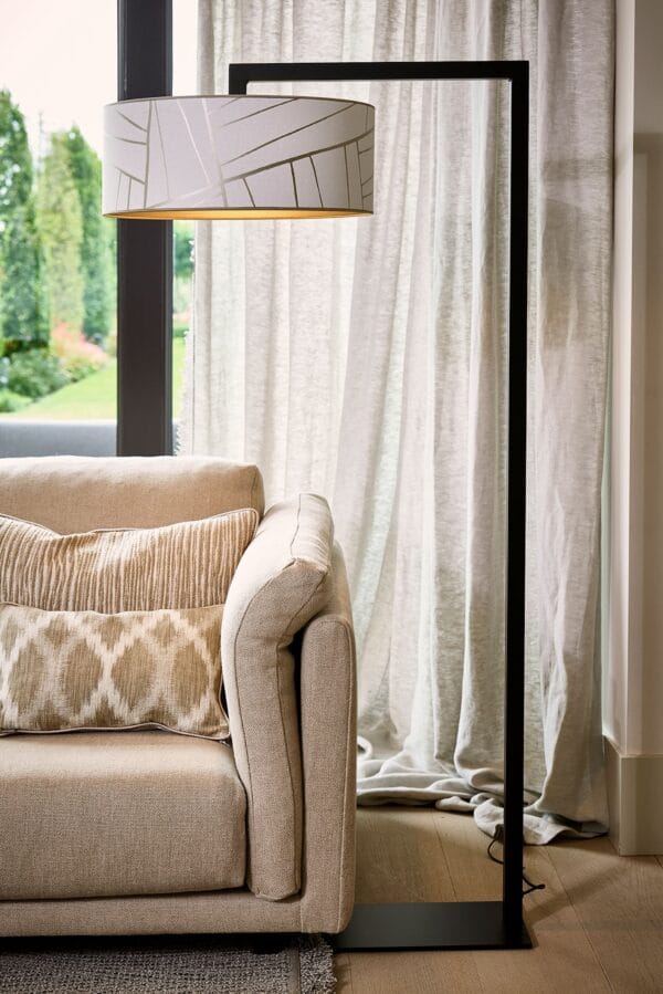 Cato floor lamp with a cylindrical light shade in a minimalist black metal frame next to a beige sofa.
