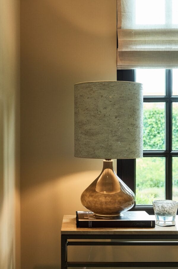Paula table lamp placed near a window, featuring a bronze reflective base and a soft grey shade.