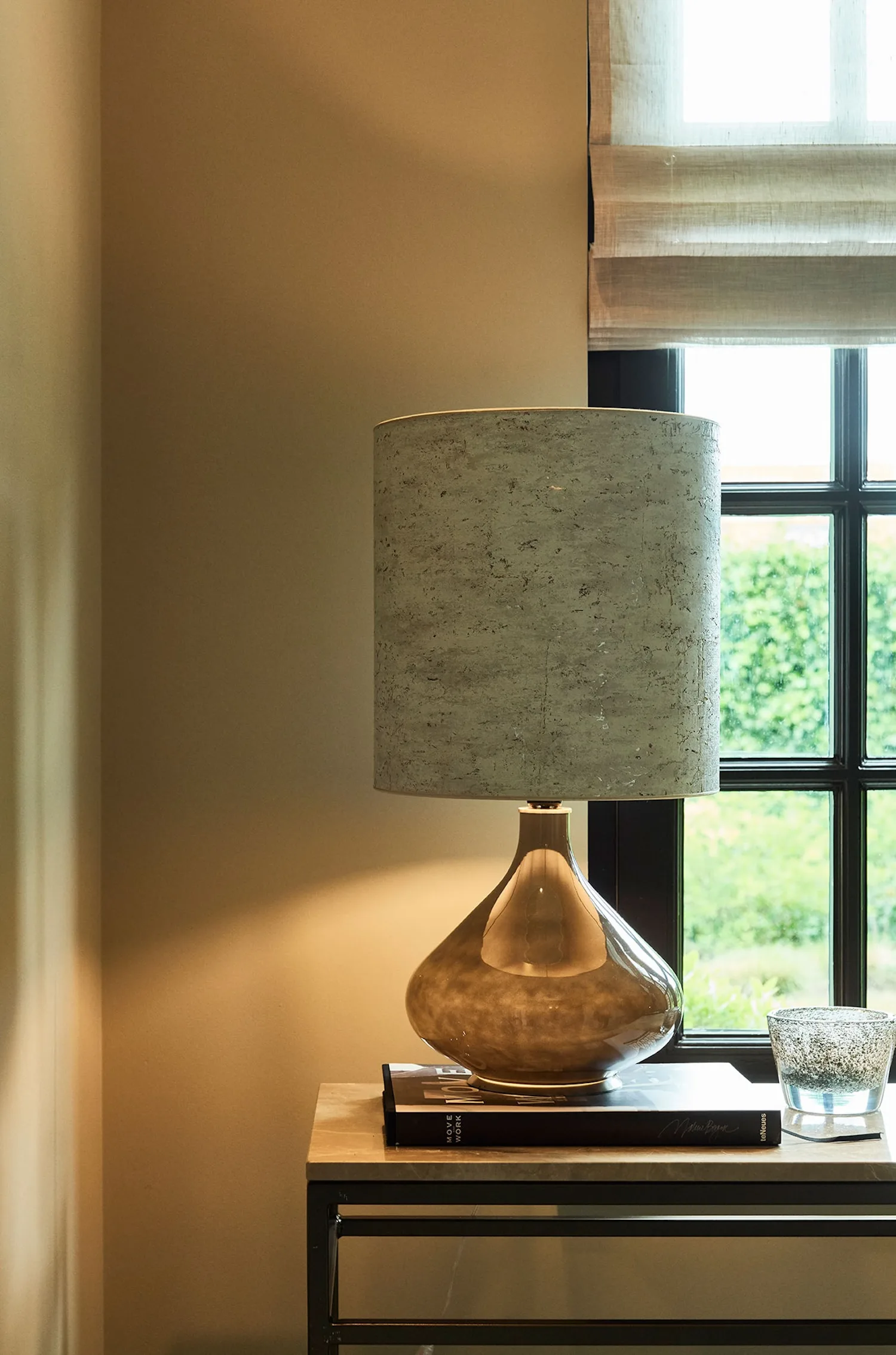 Paula table lamp placed near a window, featuring a bronze reflective base and a soft grey shade.