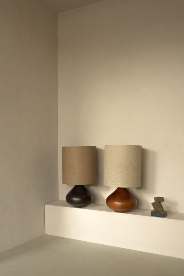 Two Paula table lamps on a ledge, one in dark wood with brown shade, one in amber wood with pale shade.