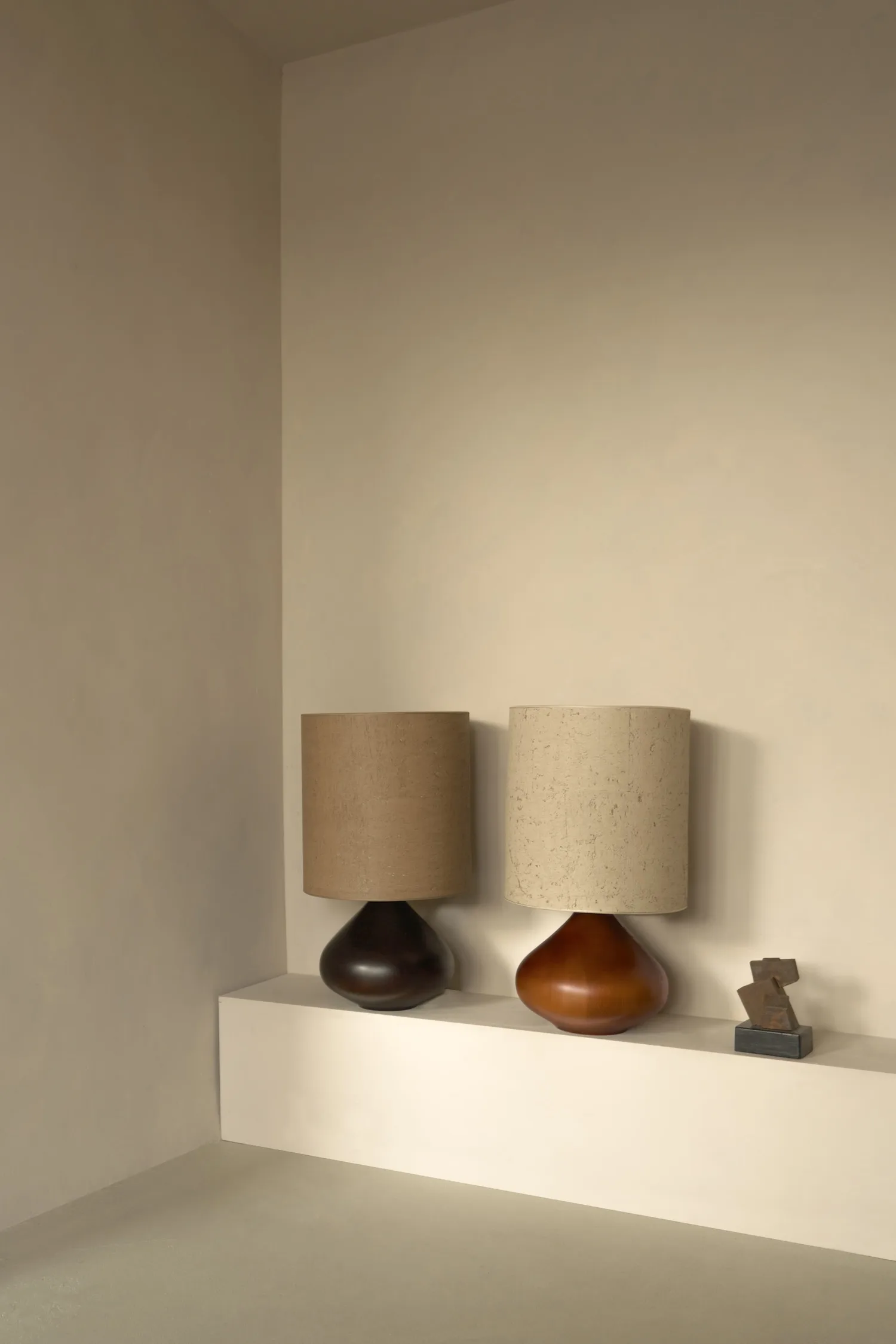Two Paula table lamps on a ledge, one in dark wood with brown shade, one in amber wood with pale shade.