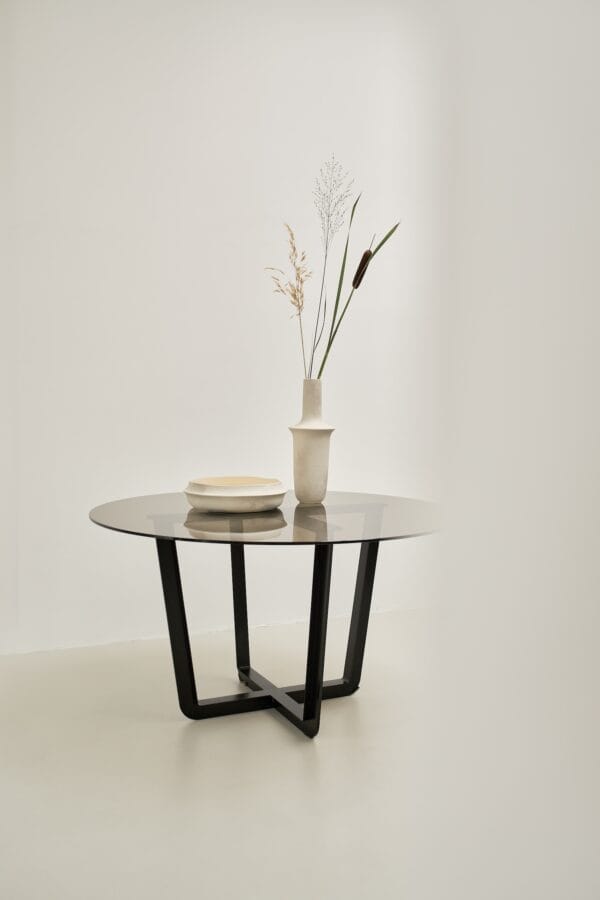 Round glass Pietro dining table with black crossed base, styled with ceramic decor and dried flowers.