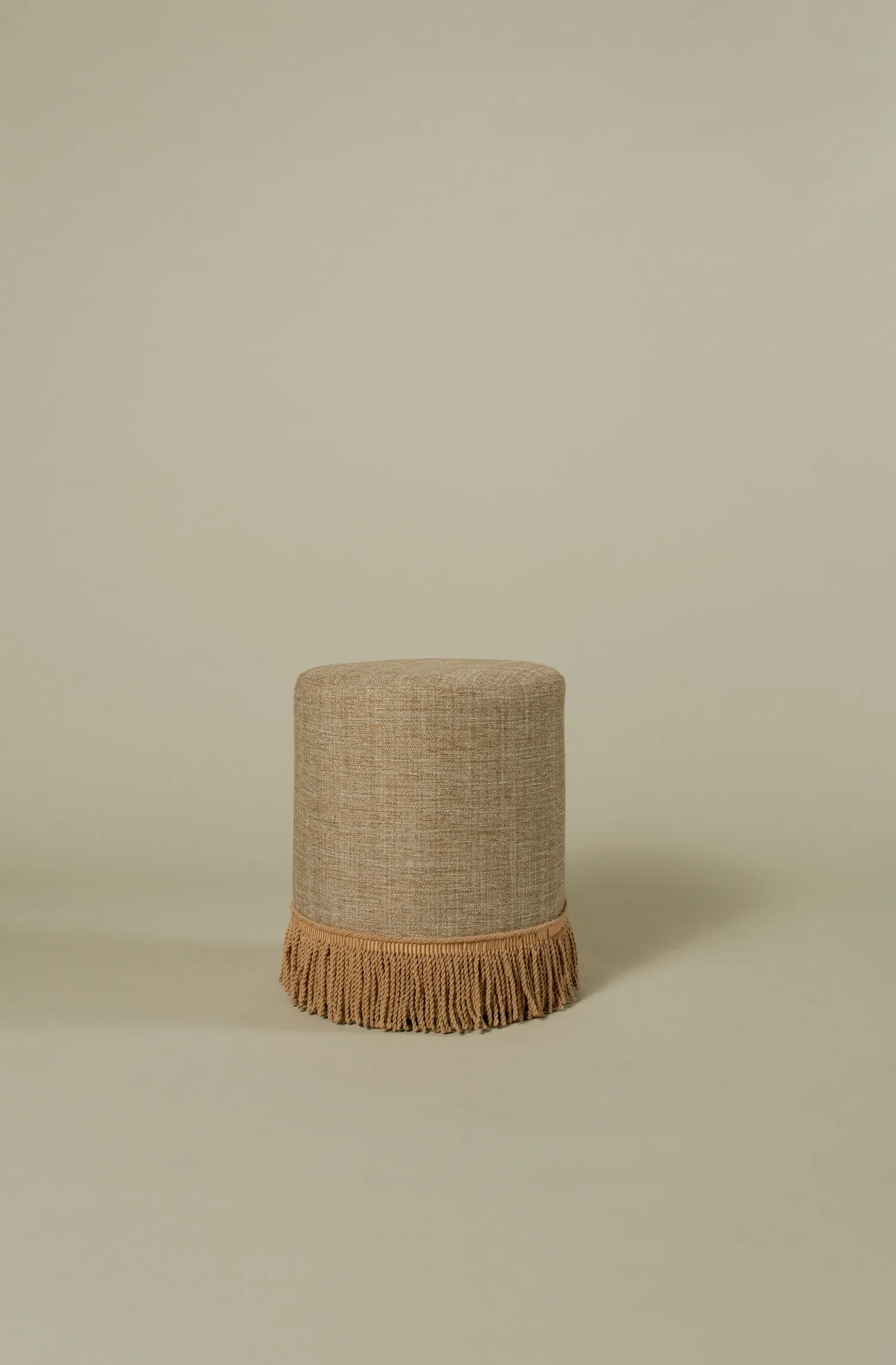 Cylindrical pouf in tan fabric, finished with a decorative fringe around the base.
