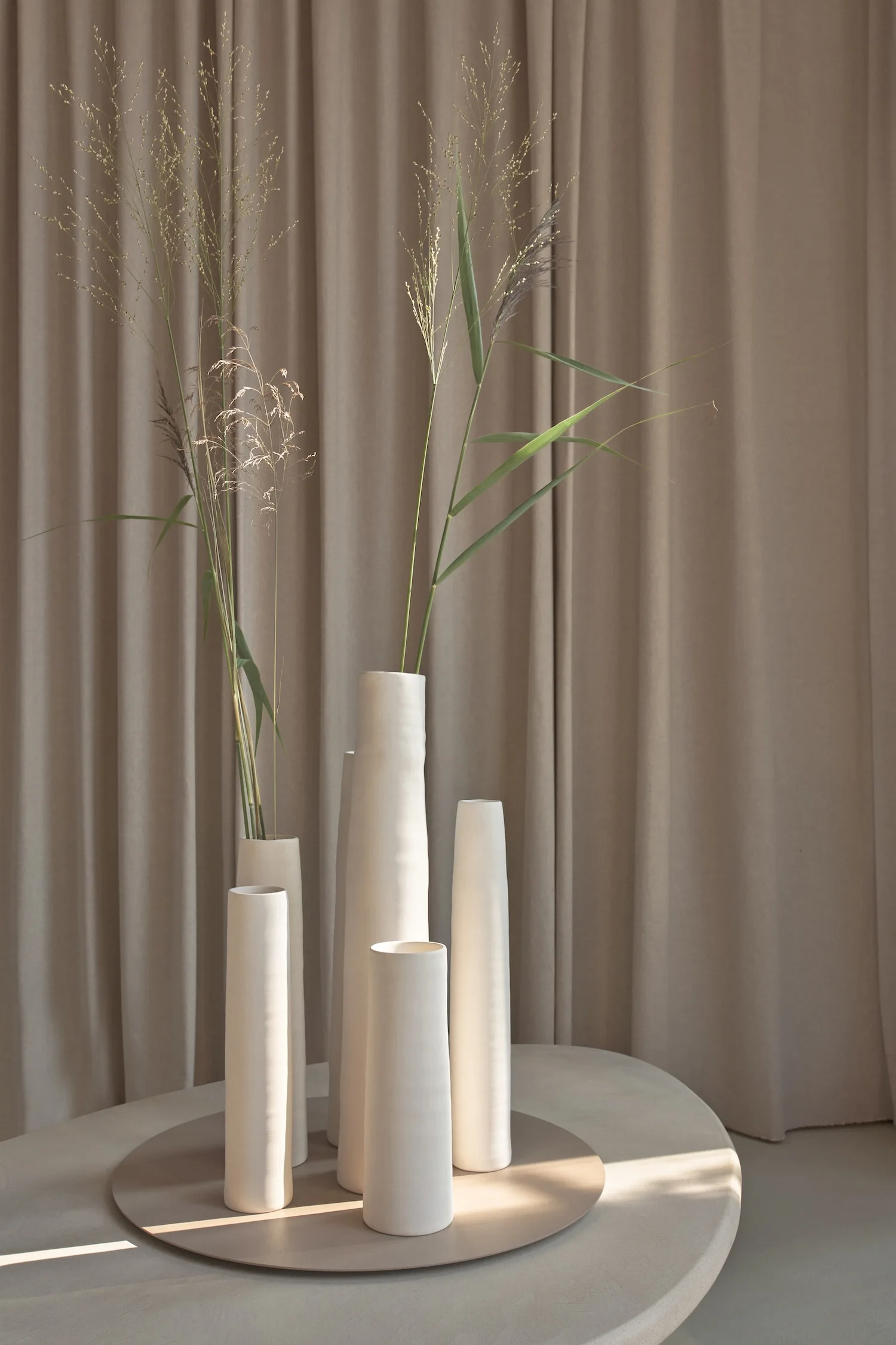 Six sculptural ceramic vases with dried grasses, placed on a round tray in front of soft drapes.