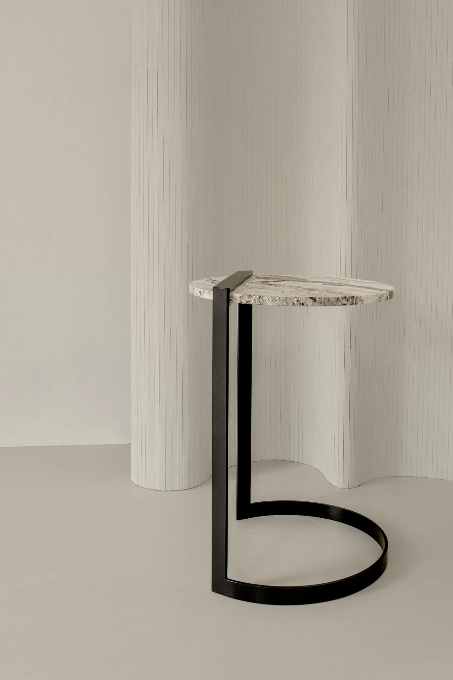 Butler side table with a light travertine top and curved black metal frame against a neutral backdrop.