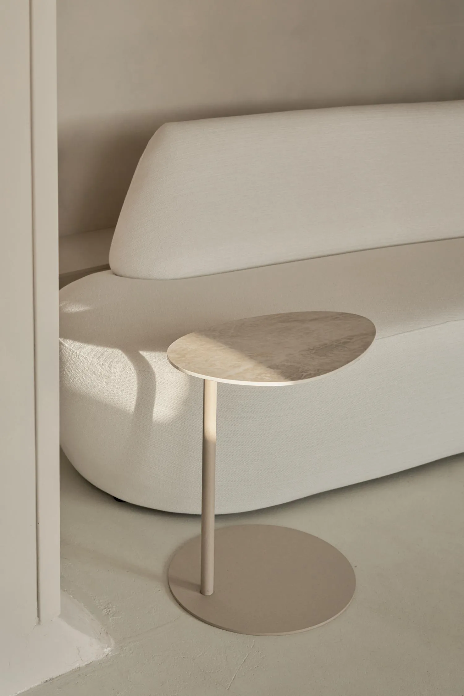 Como Butler side table with an oval travertine top and beige metal base, placed next to a soft beige sofa.