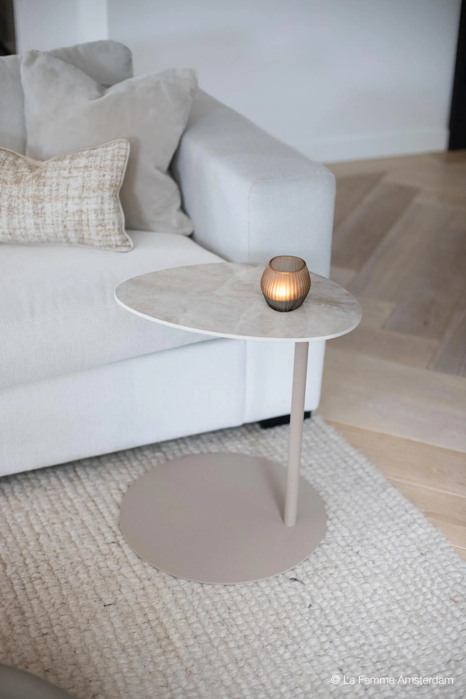 Como Butler side table with a light stone top and round metal base, styled beside a light grey sofa with a lit candle on top.