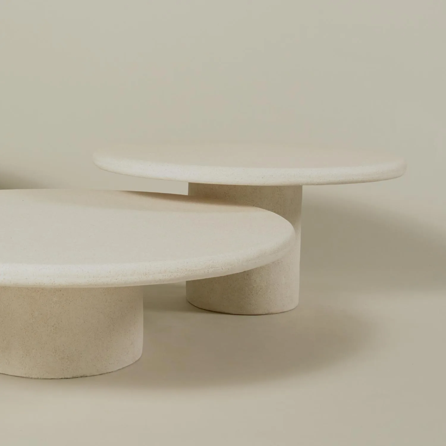 Minimalist set of two round, off-white stone coffee tables with cylindrical bases, styled against a neutral beige backdrop.