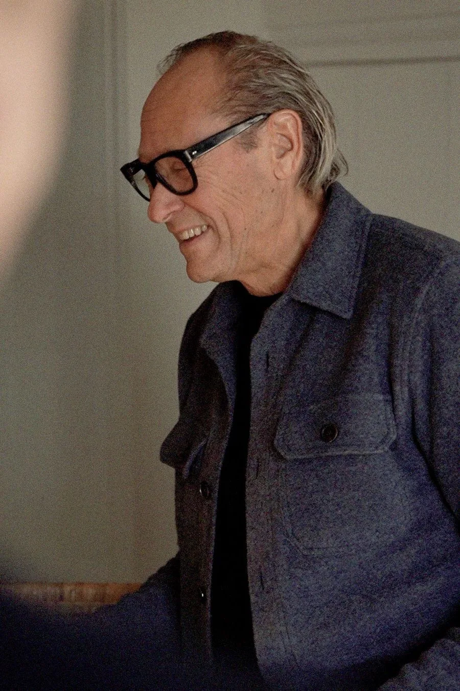 Smiling man with glasses and a gray button-up jacket, standing indoors in soft lighting.