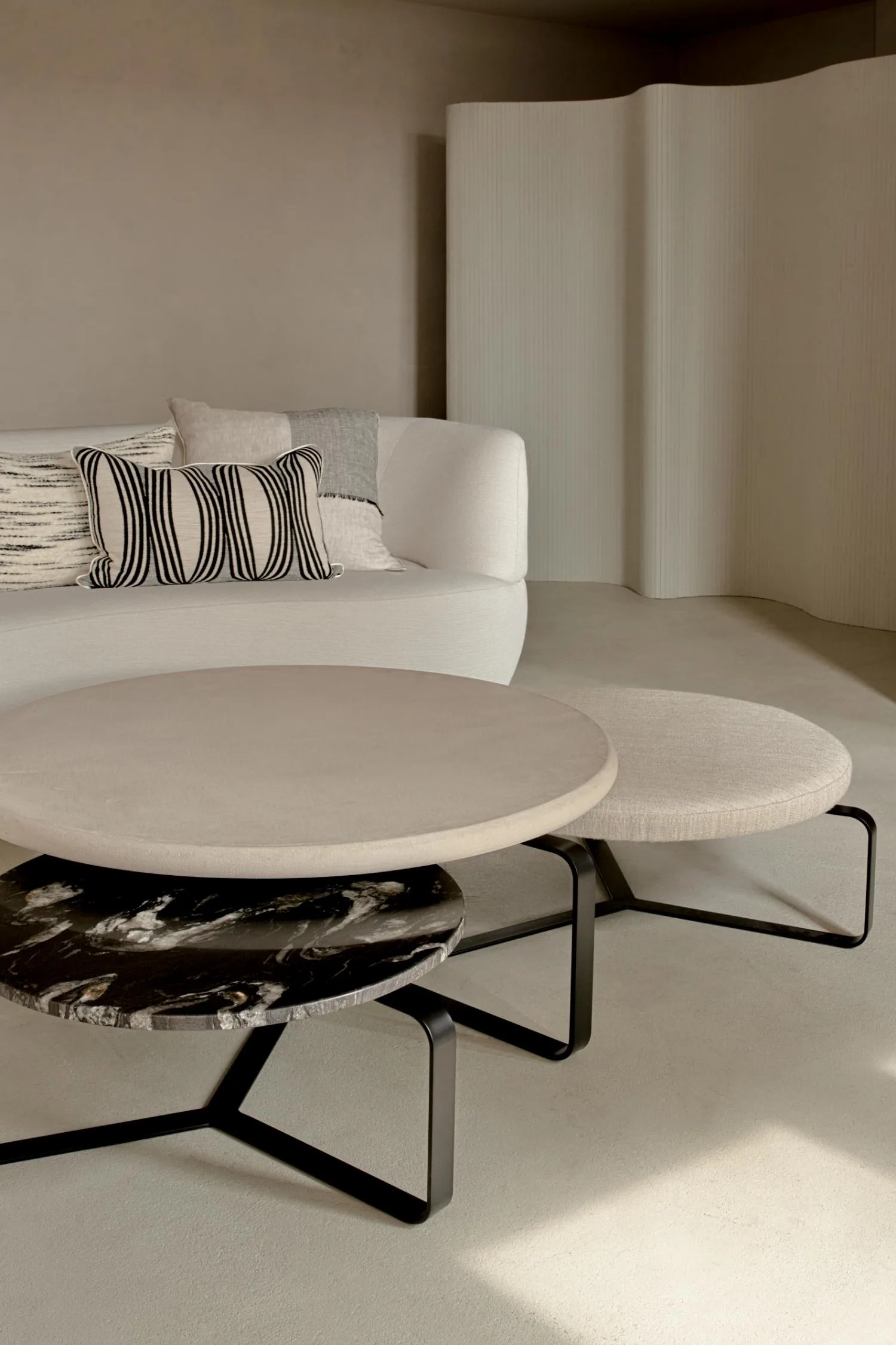 Set of three Lora coffee tables in round shapes with varying heights and finishes — beige stone, light fabric, and black marble — placed in a minimal living room.