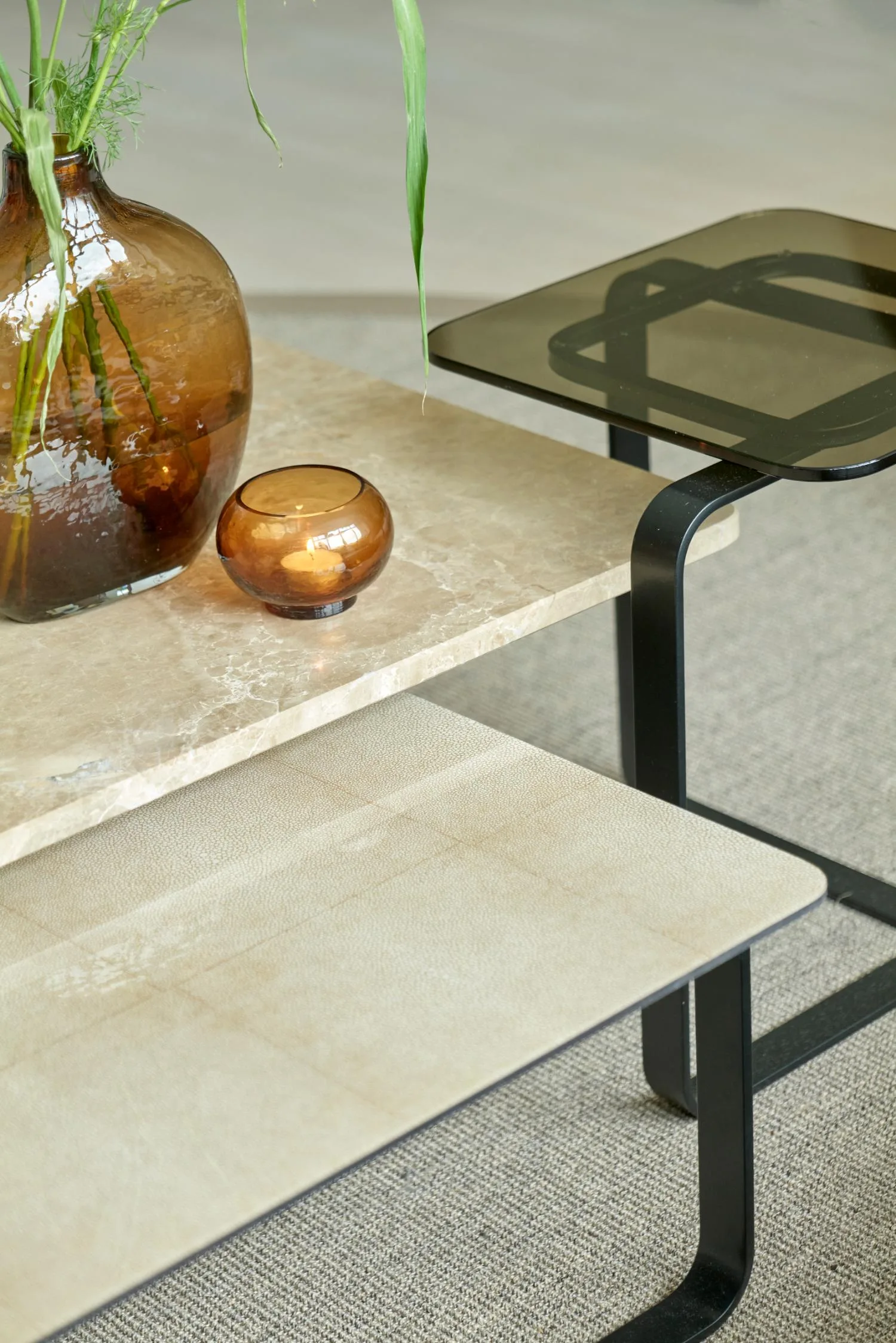 Close-up of stacked Lora coffee tables with beige stone, leather, and smoked glass tops, decorated with amber glass vases.