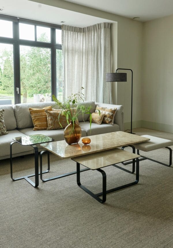 Living room featuring a complete set of Lora coffee tables with mixed materials — stone, leather, and glass — placed in front of a grey sectional sofa with patterned cushions.