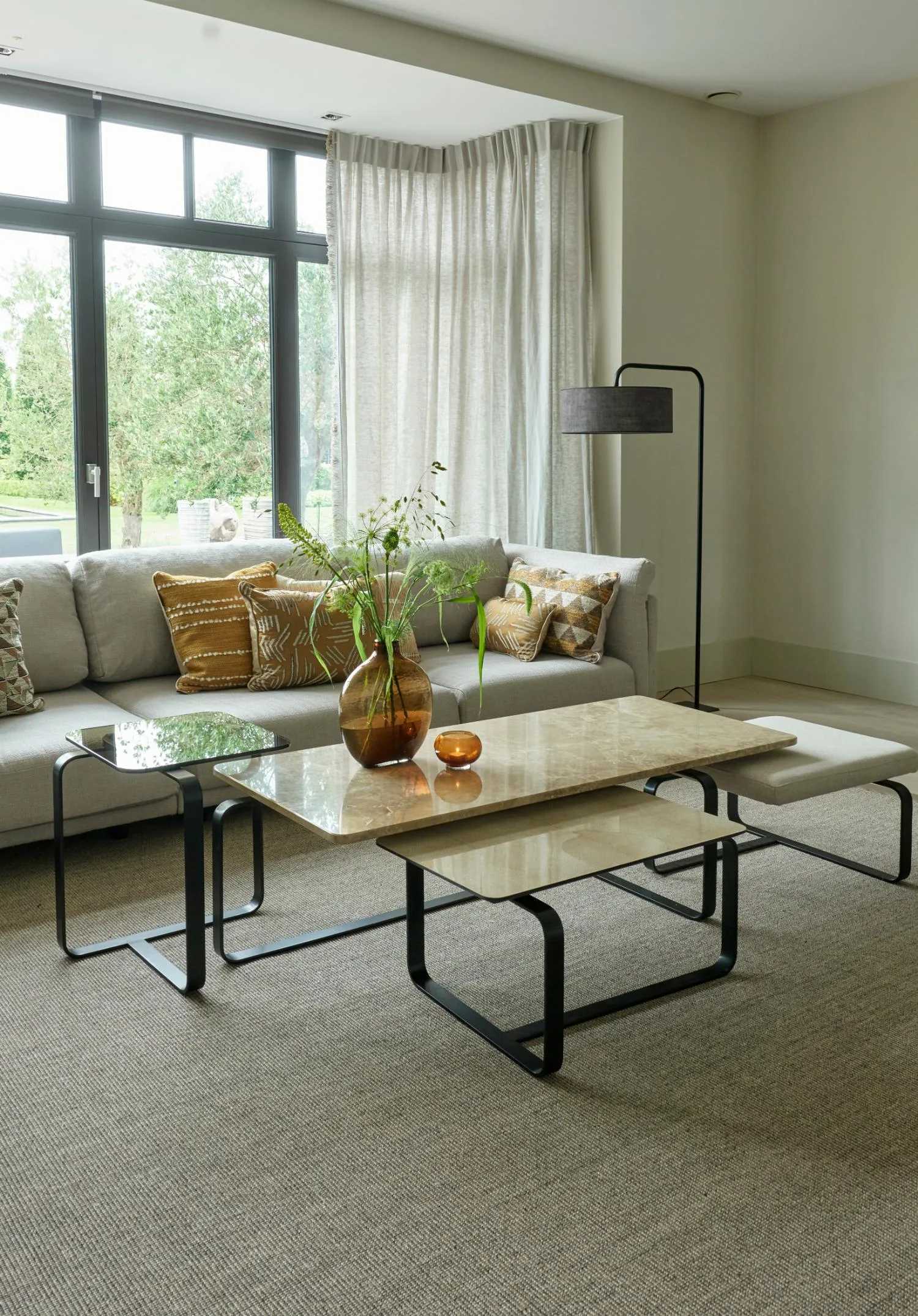 Living room featuring a complete set of Lora coffee tables with mixed materials — stone, leather, and glass — placed in front of a grey sectional sofa with patterned cushions.