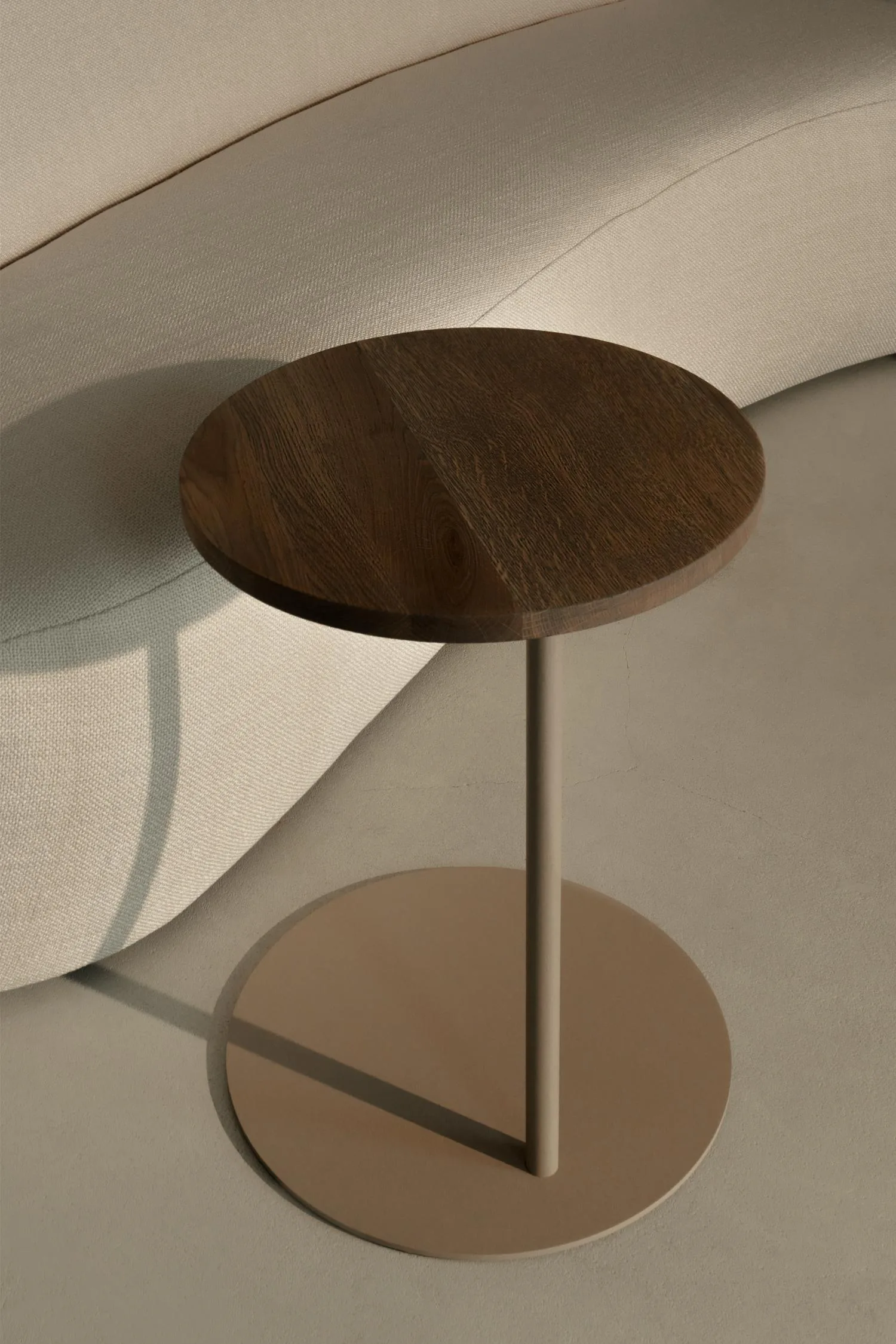 Louis Butler table with a round dark oak top and beige base, placed next to a curved light sofa.