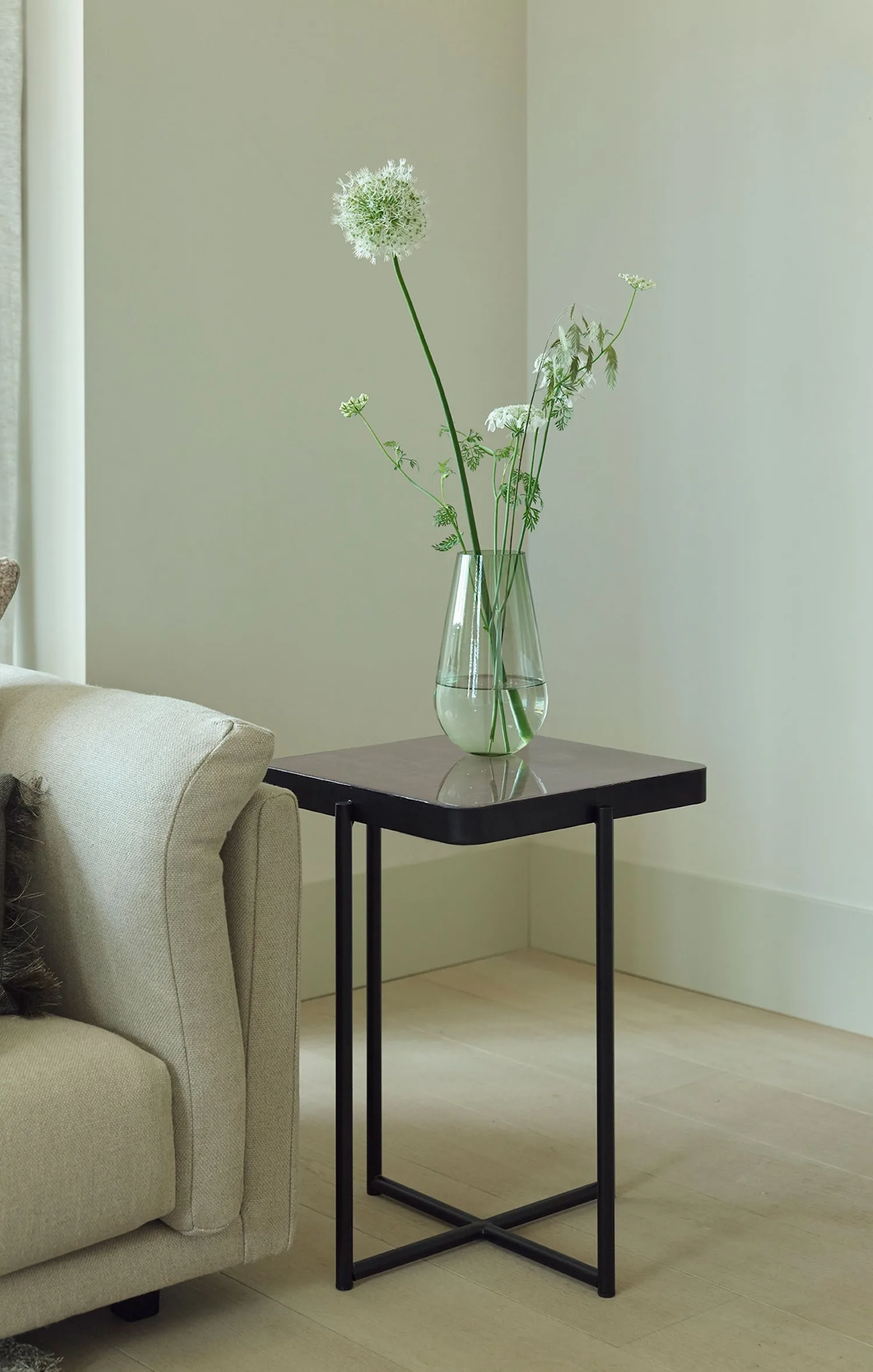 Square Lucca side table with dark glossy top and black frame, placed next to a light sofa with a glass vase of white flowers.