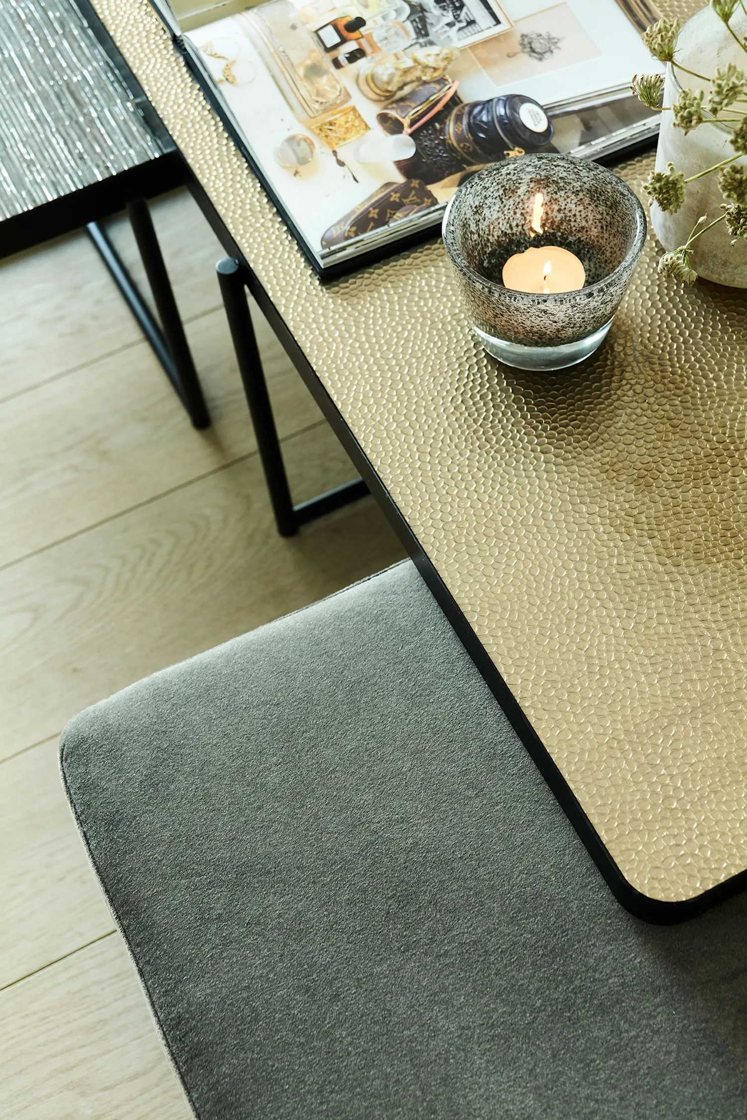 Detailed view of Lucca table with hammered gold-textured glass top, candle, and soft grey pouf below.