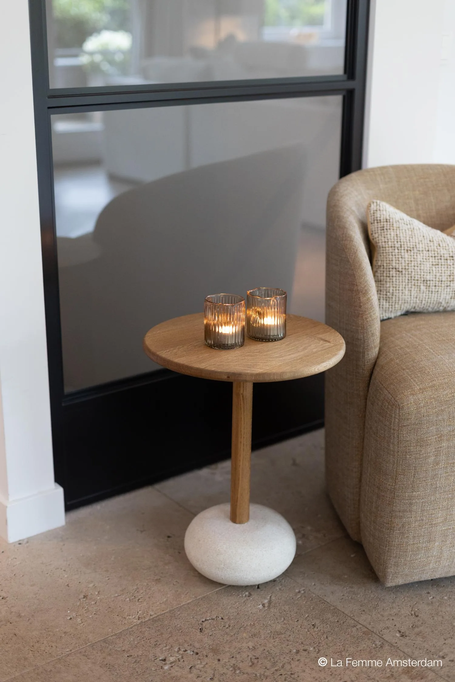 Owen side table with wooden top and stone-like round base, styled with two lit glass candle holders beside a beige armchair.