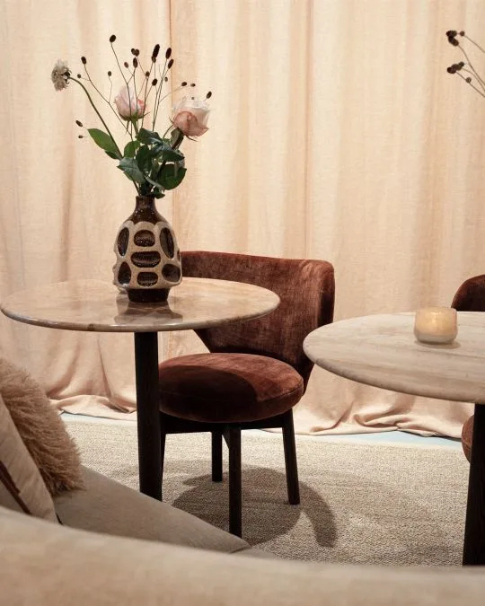 Cozy seating area featuring round marble tables, velvet dining chairs, and a ceramic vase with fresh flowers against a beige curtain backdrop.
