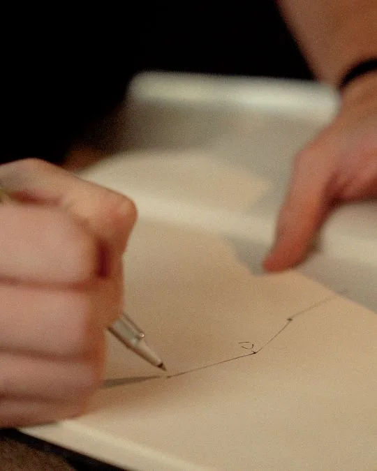 Close-up of a hand sketching a geometric line drawing with a pen on a blank notebook page.