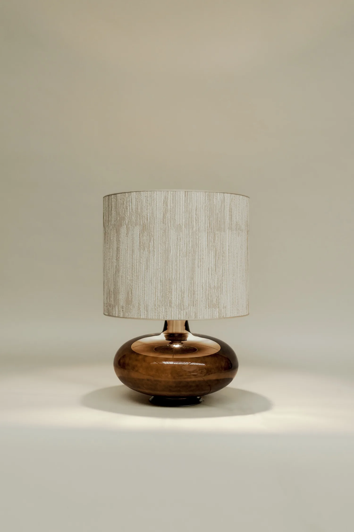 Katie table lamp with a round amber glass base and textured beige lampshade, set against a neutral background.