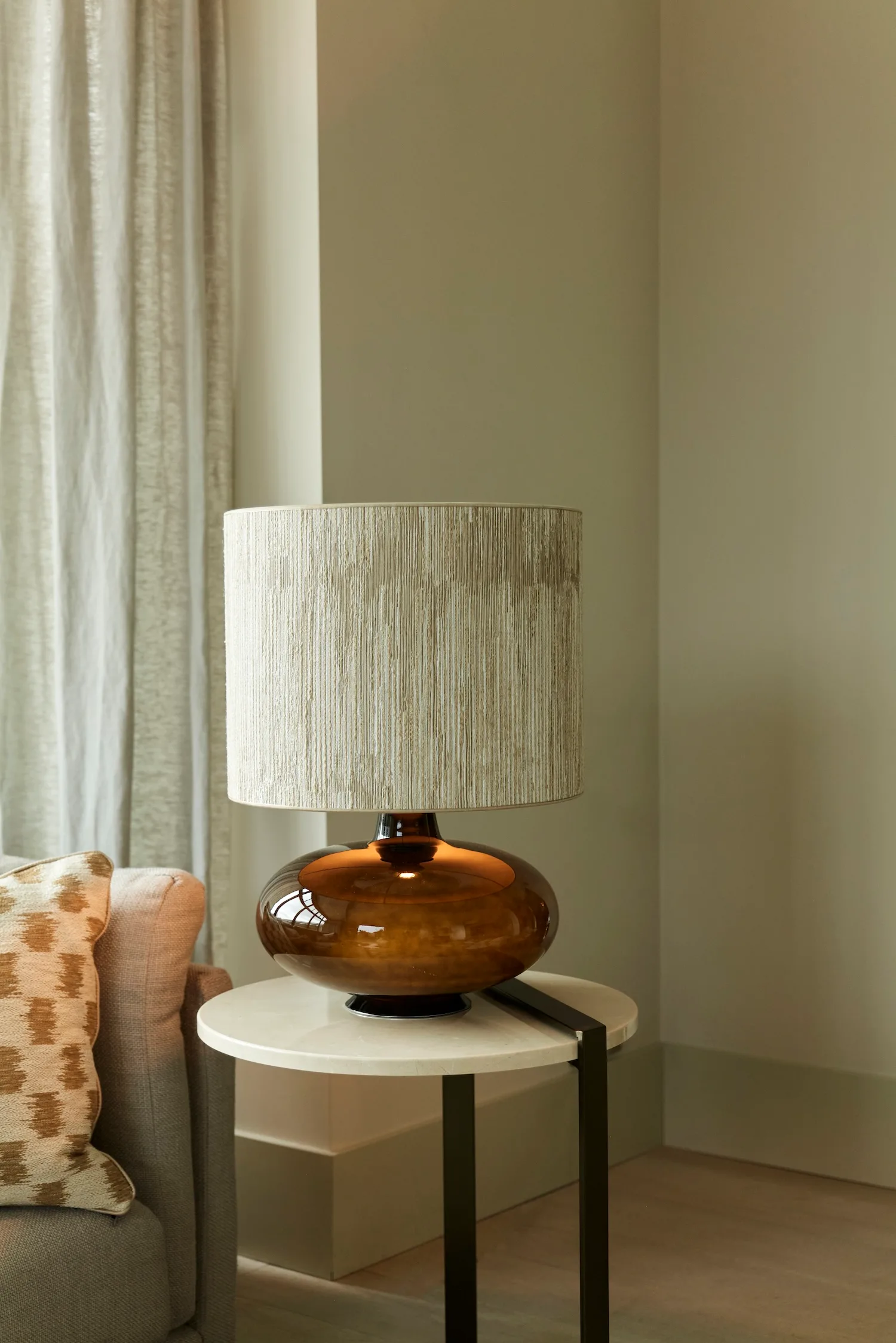 Katie table lamp with amber glass base and neutral textured shade, styled on a round marble side table next to a beige sofa.