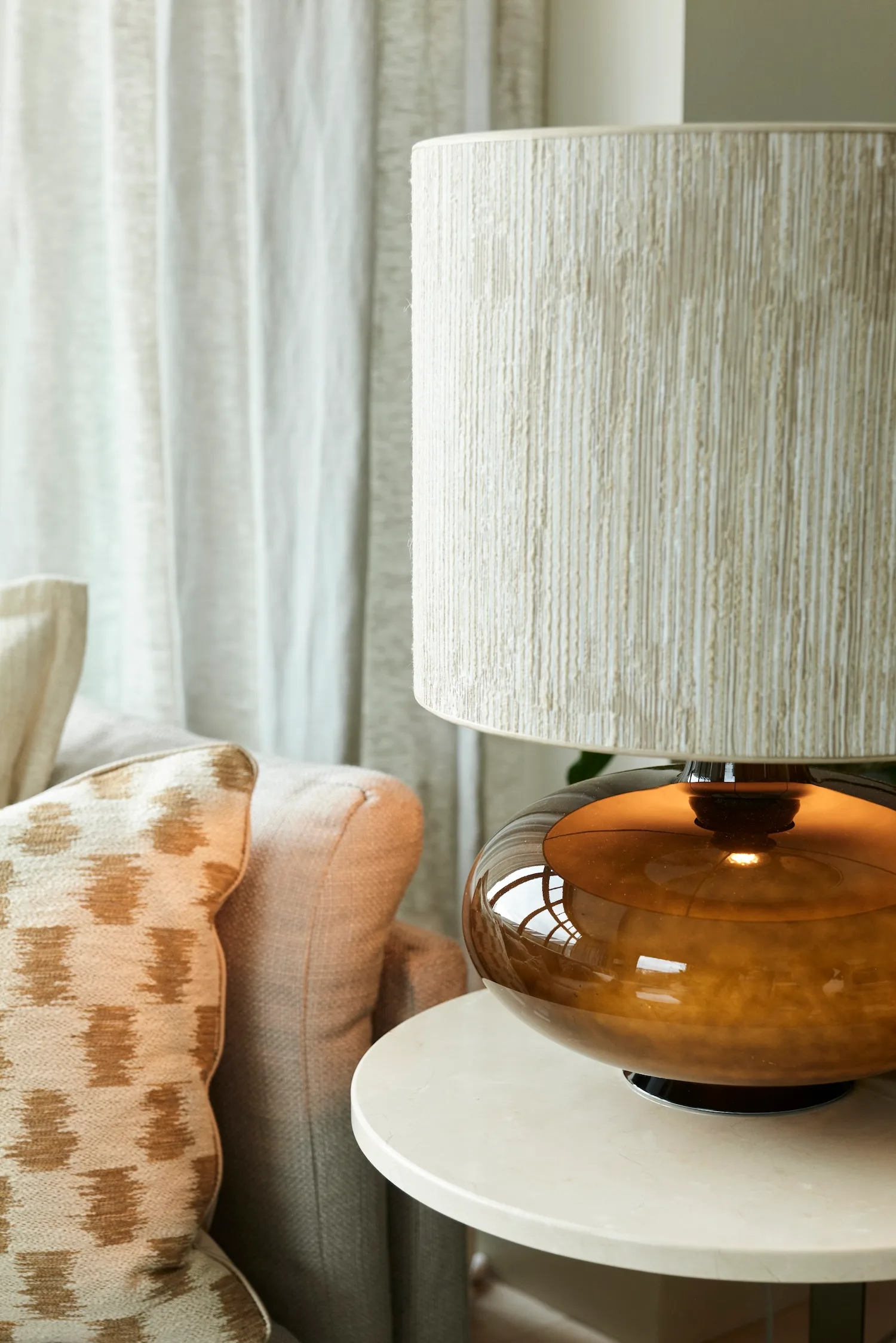 Close-up of the Katie table lamp with a rounded amber glass base and textured neutral lampshade, placed on a round marble side table next to a beige sofa.